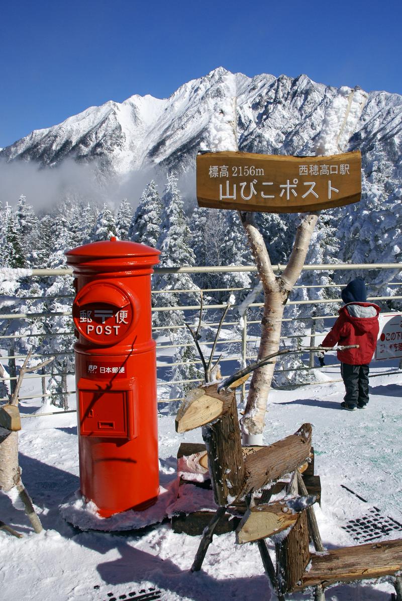 紅色的日本郵便圓筒郵筒立在雪地裡,旁邊的木牌寫著「標高2156m 西穂高口駅 山びこポスト」,背後是覆雪的西穗高岳與白樺欄杆