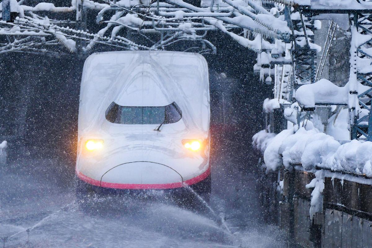 Head-on view of a Joetsu Shinkansen train running into Echigo-Yuzawa Station through heavy falling snow, headlights on, with trackside snow-melting sprinklers spraying water and thick snow piled on the catenary supports and lineside fence