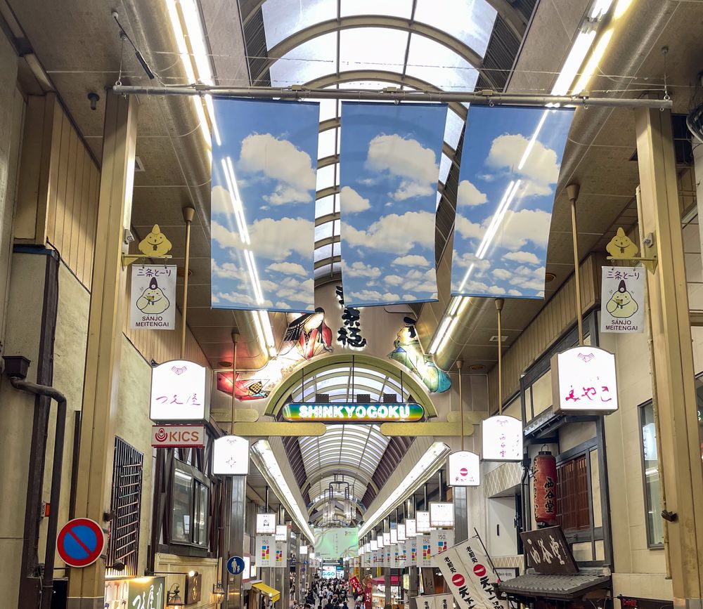 新京極商店街的拱廊入口:玻璃拱頂下掛著三幅藍天白雲圖案的布幔,中央拱門上有彩色的 SHINKYOGOKU 字樣,兩側柱子掛著「三条とーり SANJO MEITENGAI」的旗子與店家燈箱,拱廊往深處延伸