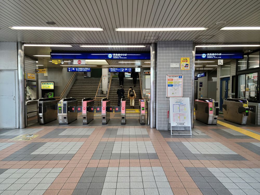 京急汐入駅的剪票口,上方藍色看板寫京急線のりば,閘門後方兩道樓梯,指示牌分別標 1 號與往橫濱・品川方面的 2 號月台