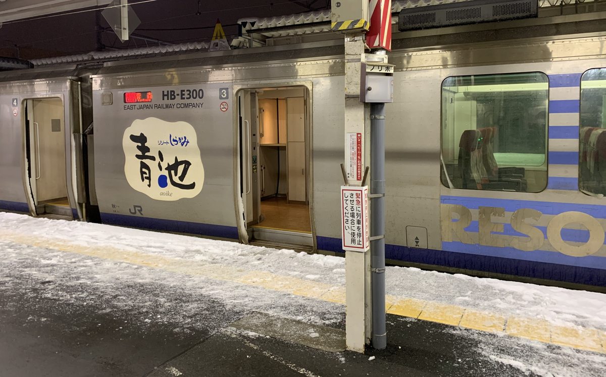 夜晚的青森站月台積著雪，停著 Resort 白神號青池編成：銀色車身上印著「HB-E300」「3」號車與圓形的「青池」標誌，車門開著，旁邊車廂側面有藍白條紋和「RESORT」大字
