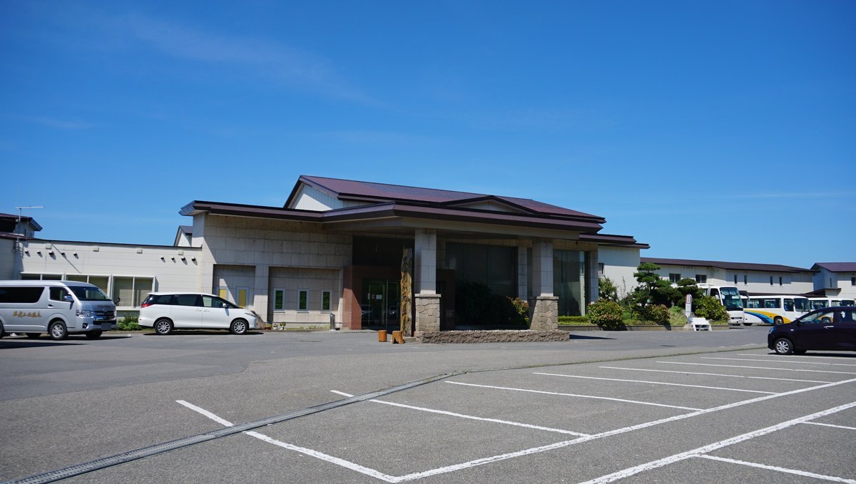 黄金崎不老ふ死温泉的旅館正門：一棟紅褐色屋頂的低矮建築，入口有石柱門廊，前面是空曠的停車場，左邊停著一台車身用金字印著「不老ふ死温泉」的銀色廂型車，右後方停著兩台大型遊覽巴士