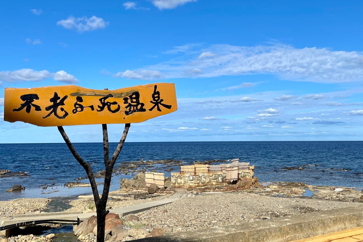 海邊步道旁一塊橘黃色木牌，手寫毛筆字「不老ふ死温泉」，牌子後方的礁岩海岸上，有一圈用石頭與木板圍起來的露天浴池圍欄，再往外是藍色的日本海