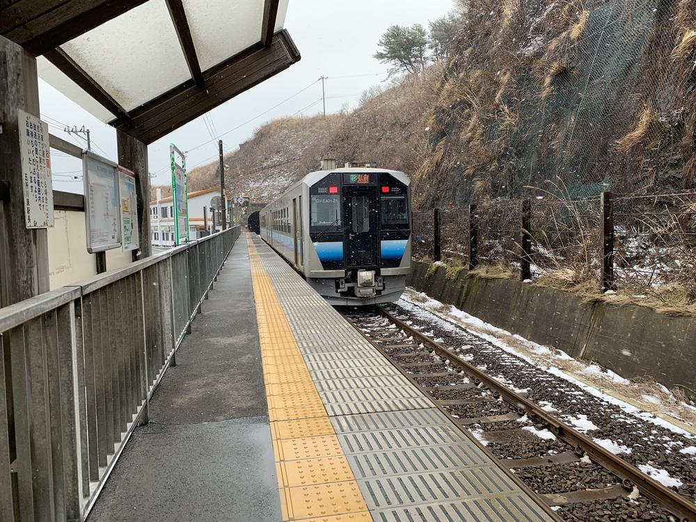 下著小雪的千疊敷站單線月台，一列銀色車身、藍色腰帶的 GV-E400 系普通車停在月台旁，車頭顯示「弘前」，右邊是覆著乾草與防落石網的山壁，左邊月台雨棚下貼著告示
