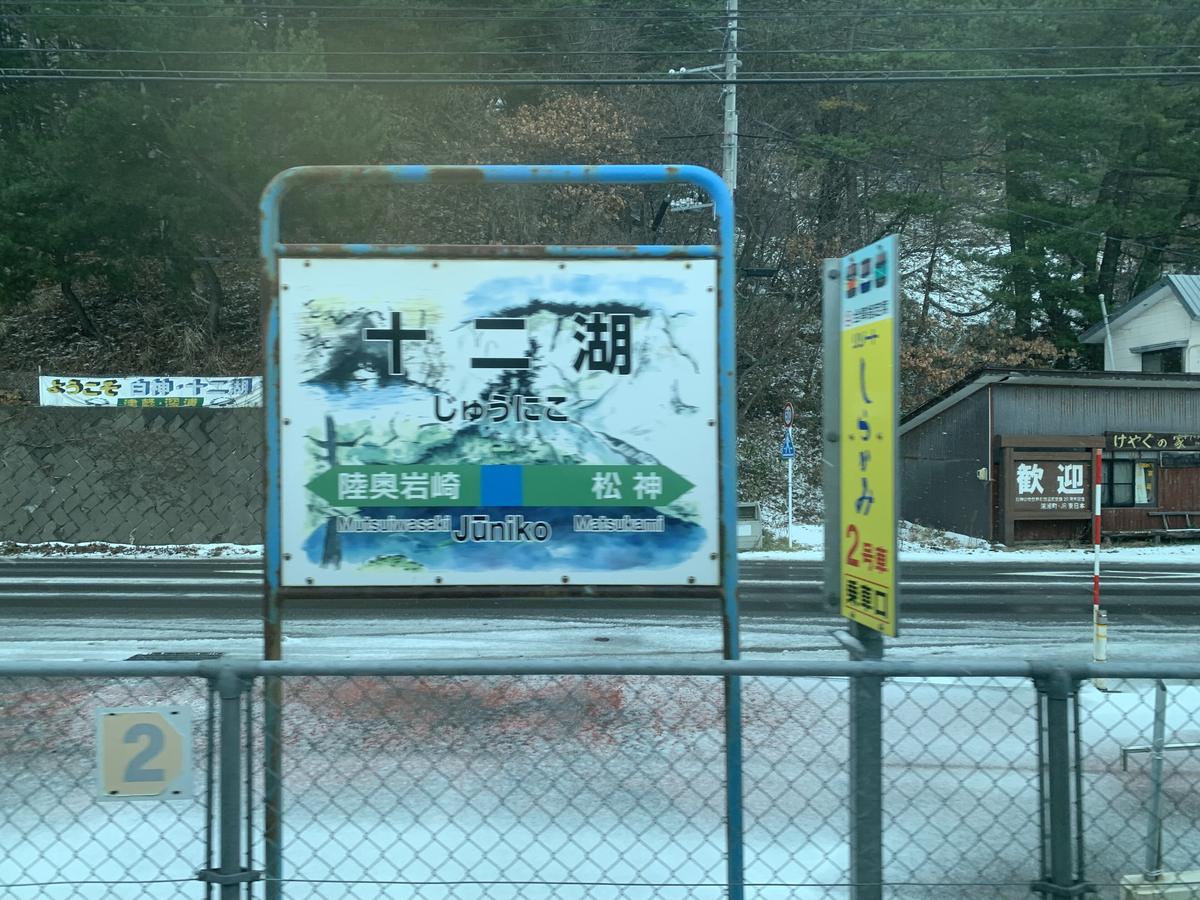 從車窗看出去的十二湖站月台：藍框站名牌寫「十二湖 じゅうにこ」，左右箭頭是陸奧岩崎與松神，旁邊一塊黃色立牌寫「リゾートしらかみ 2号車 乗車口」，地上有薄雪，遠處是「ようこそ 白神・十二湖」的布條與寫著「歓迎」的小屋