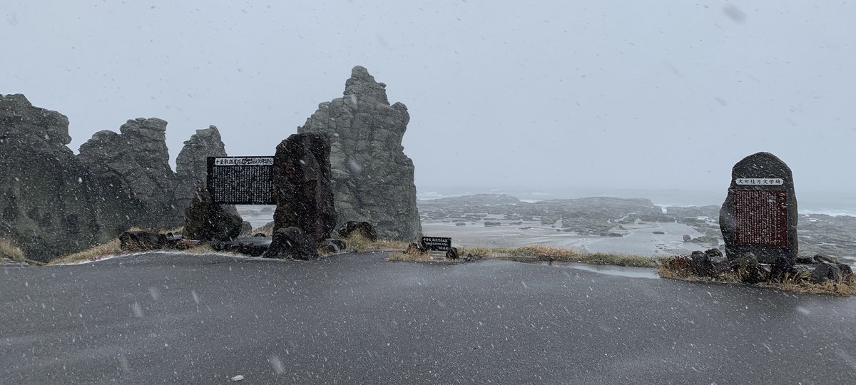 大雪中的千疊敷海岸：前景是濕的柏油廣場，左邊幾塊高聳的灰色奇岩與刻字的石碑，右邊一塊「大町桂月文学碑」，後方是平坦的岩床一路延伸到起浪的海
