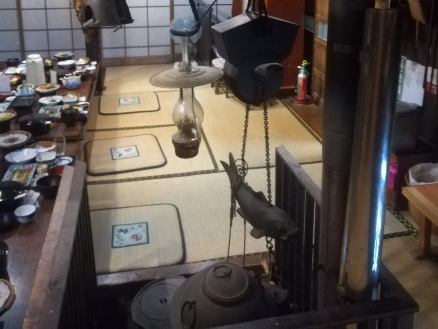 The hearth room of a gassho-zukuri guesthouse: a sunken irori hearth in the foreground with a large iron kettle, a chain and a carved fish-shaped pot hook hanging above it; along the left a long low table laid with individual place settings of small bowls, black lacquer soup bowls, blue-and-white dishes and a teapot, three flat seat cushions in a row on the tatami, shoji screens and soot-blackened posts behind