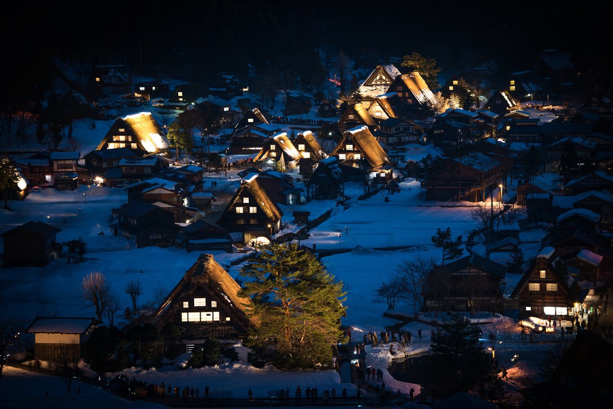 Ogimachi gassho village during the Shirakawa-go illumination: floodlights turn the snow-laden thatched roofs gold while crowds line the village lanes and viewing area