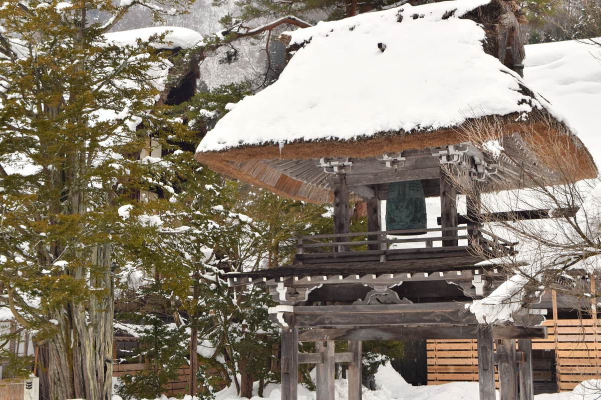 Myozenji's two-tier thatched bell-tower gate under snow: a verdigris bronze bell hangs in the open upper chamber behind a wooden railing, the thatched roof carries a thick cap of snow with icicles along the eave, carved brackets on the lower gate tier, a snow-laden cedar at left, snow-covered gassho thatch and a wooden plank fence behind, deep snow on the ground, nobody in frame