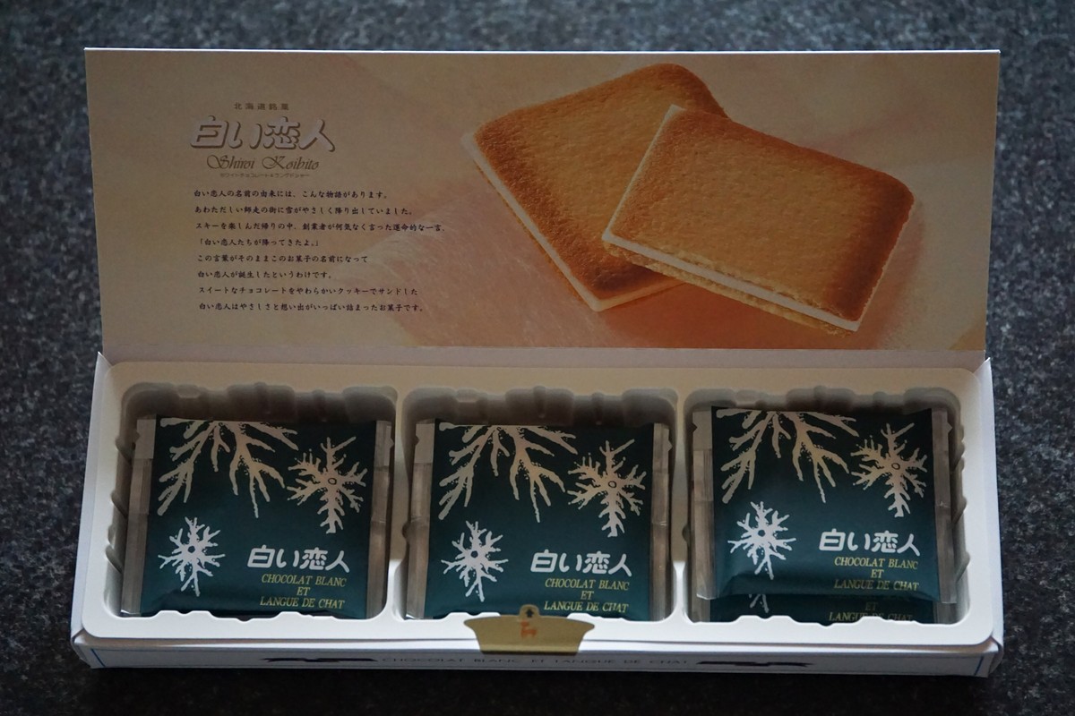 打開的白色戀人紙盒:上蓋內側印著兩片夾白巧克力的長方形貓舌餅乾照片與日文由來說明,下層整齊排著三個深綠色的獨立包裝,包裝上印著白色雪花與「白い恋人 CHOCOLAT BLANC ET LANGUE DE CHAT」