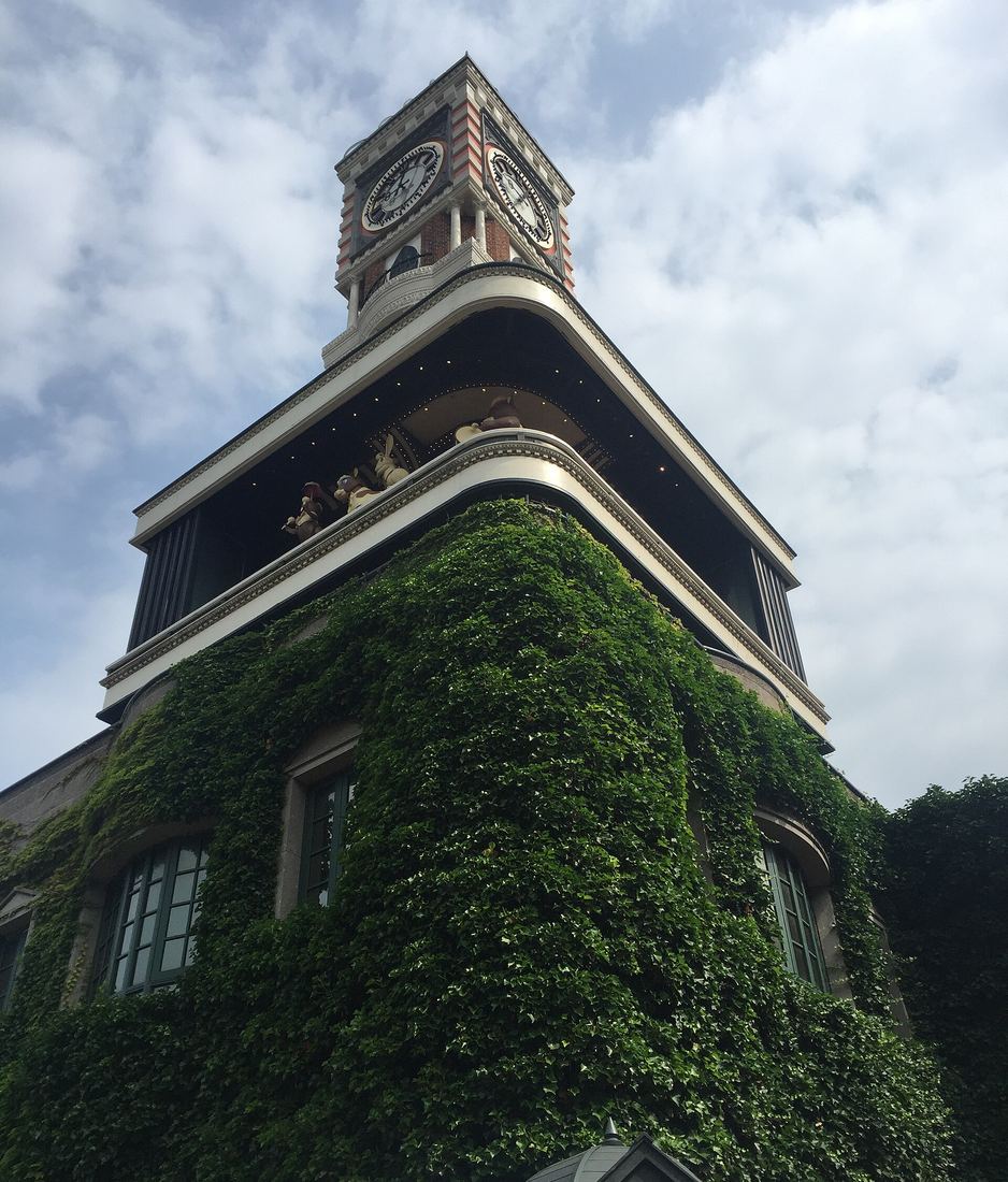 從正下方仰看白色戀人公園的機關鐘塔:塔頂兩面圓鐘,下一層的陽台欄杆後方探出好幾隻米白與粉紅色的人偶,塔身牆面爬滿常春藤