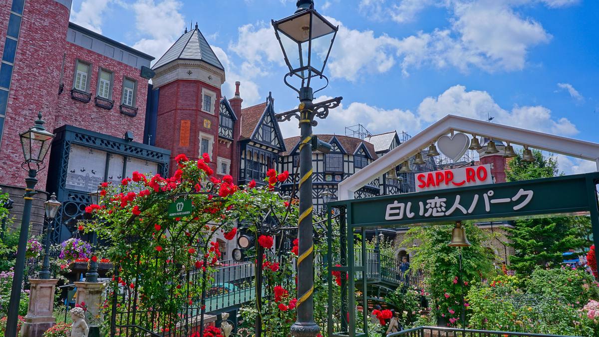 白色戀人公園玫瑰花園裡的綠色鐵製招牌拱門,上面寫著白色的「白い恋人パーク」和紅色的「SAPPORO」,頂端掛著一排銅鈴與一顆白色愛心,前景是攀著紅玫瑰的鐵架與古典街燈,背景是半木造的英式樓房