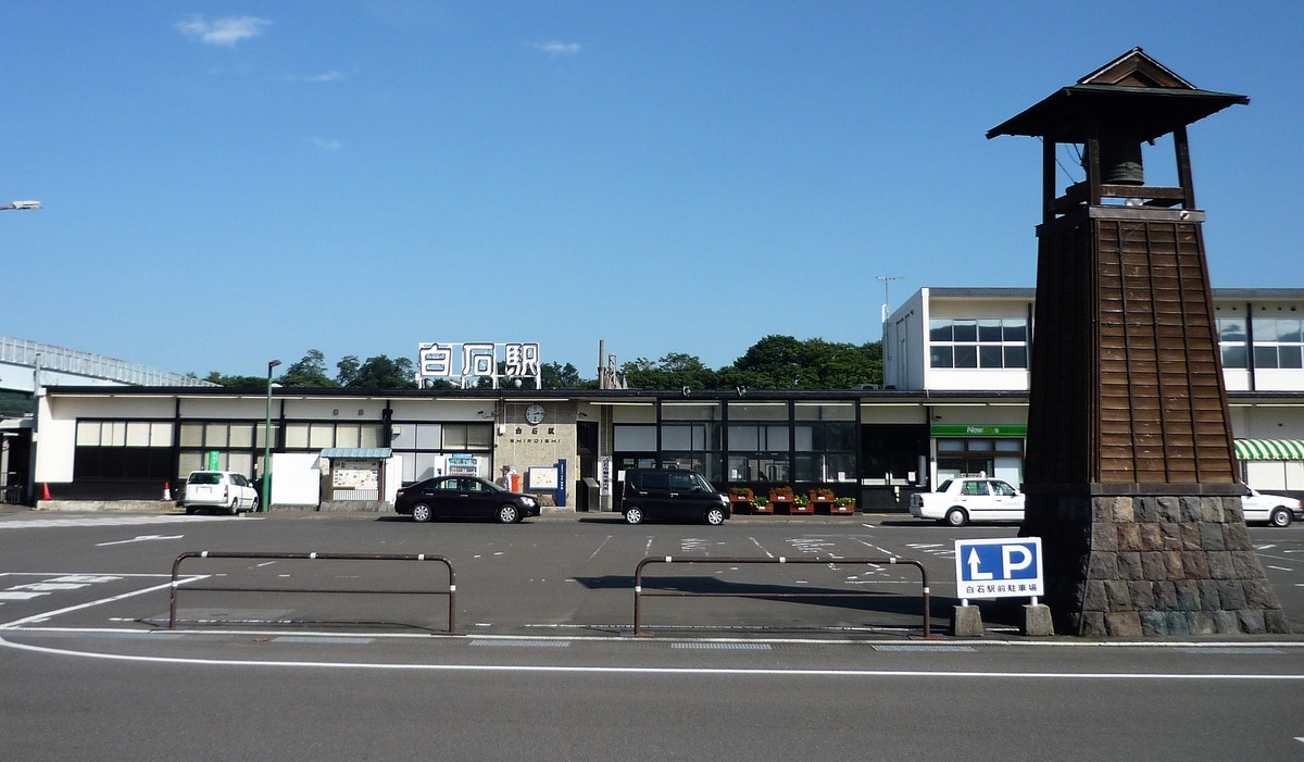 JR 白石駅站舍,白色屋頂上立著「白石駅」三個大字,站前是柏油停車場、幾台停著的計程車與一塊站前停車場指示牌,右側一座木造鐘樓,藍天無雲