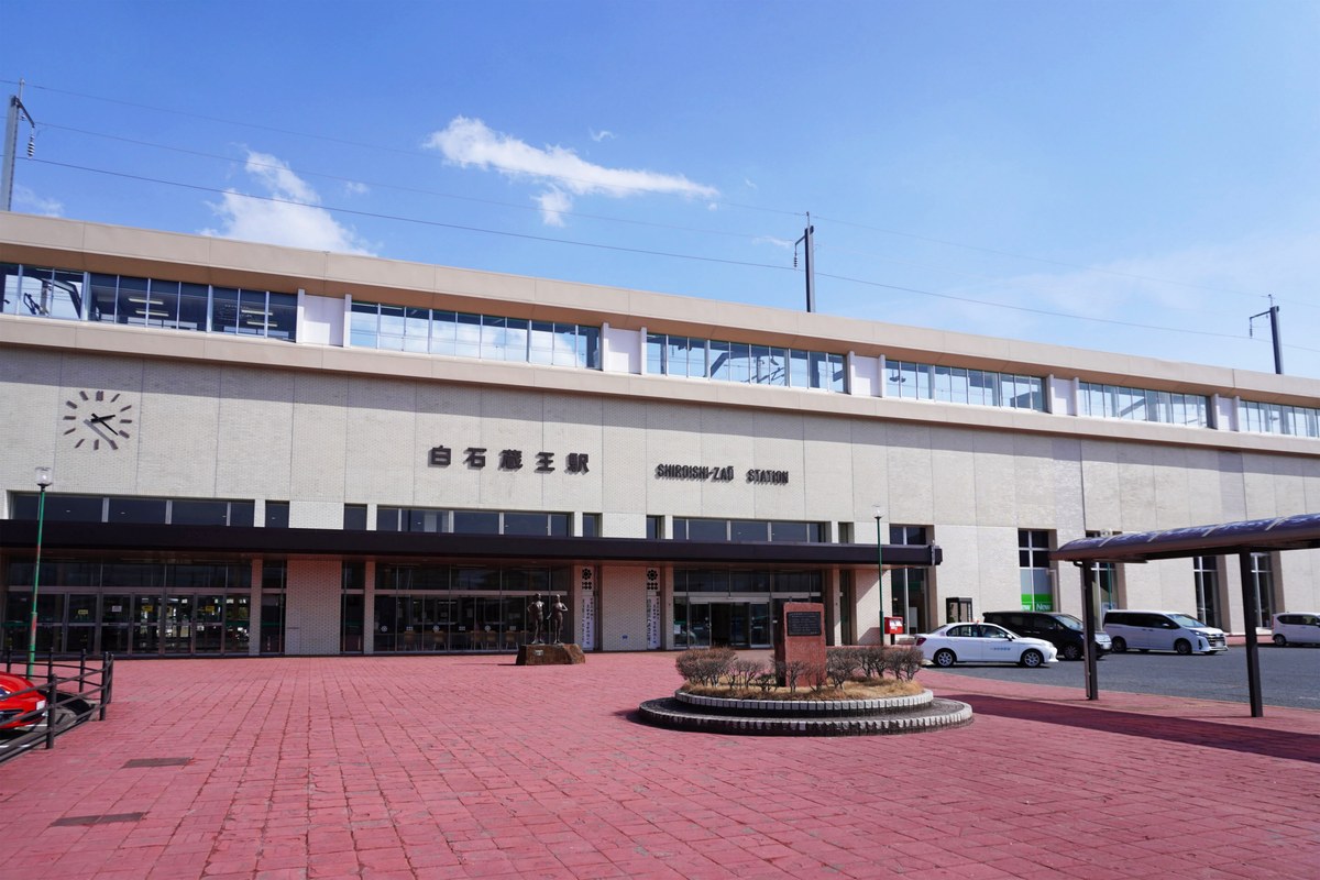 白石蔵王駅西口的站前廣場:米色磁磚外牆上有「白石蔵王駅 SHIROISHI-ZAO STATION」字樣與一面圓形時鐘,玻璃門前立著一座銅像,地面是紅磚鋪面,右側停著幾台車