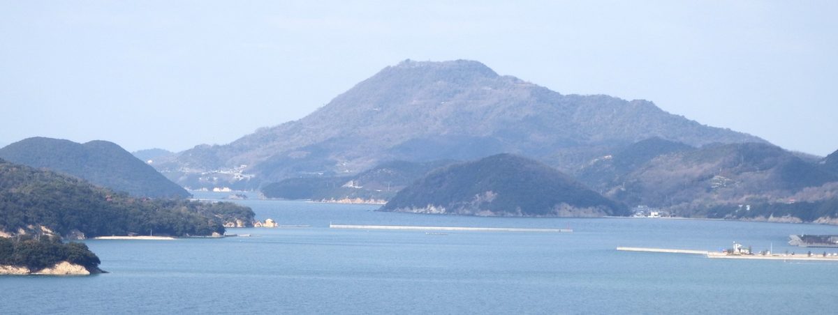 隔著平靜的海面從東南方遠望紫雲出山：中央是一座山頂略平的冬季褐綠色山，前方海上有兩座低矮的綠色小島，海面上有兩道長長的防波堤