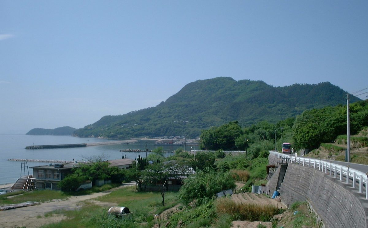 從海岸道路往西北看紫雲出山：右側是有白色護欄的彎道，一台紅色巴士正開過來，左側是低矮的房舍、海邊與伸進海裡的防波堤，後方是長滿綠樹的紫雲出山