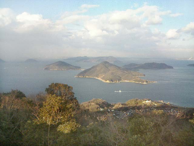 冬天從紫雲出山山頂往東北看：前景是枯黃與深綠的樹叢，山腳海邊有一小片聚落，海上有一座三角形的島與幾座低矮的島嶼，一艘白色小船正駛過，天上有大片白雲
