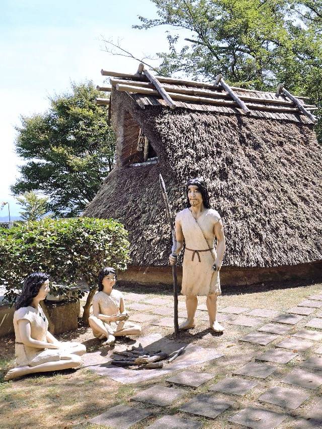 紫雲出山山頂遺跡區的復原茅草屋：一座屋頂蓋到地面的茅草豎穴住居前，站著一尊手持長矛、穿麻布衣的彌生人模型，旁邊有兩尊坐在地上的模型，地上鋪著石板