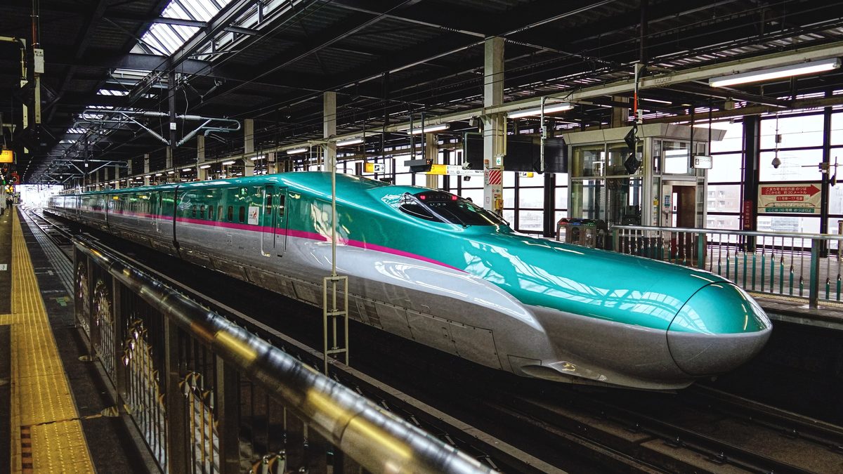盛岡站的新幹線月台上停著一列 E5 系「はやぶさ」，綠色車頭配白色車身與粉紅色腰帶，長長的車鼻伸向畫面右側，月台頂棚是深色鋼架，右側窗邊掛著往秋田方面的こまち號乘車位置看板