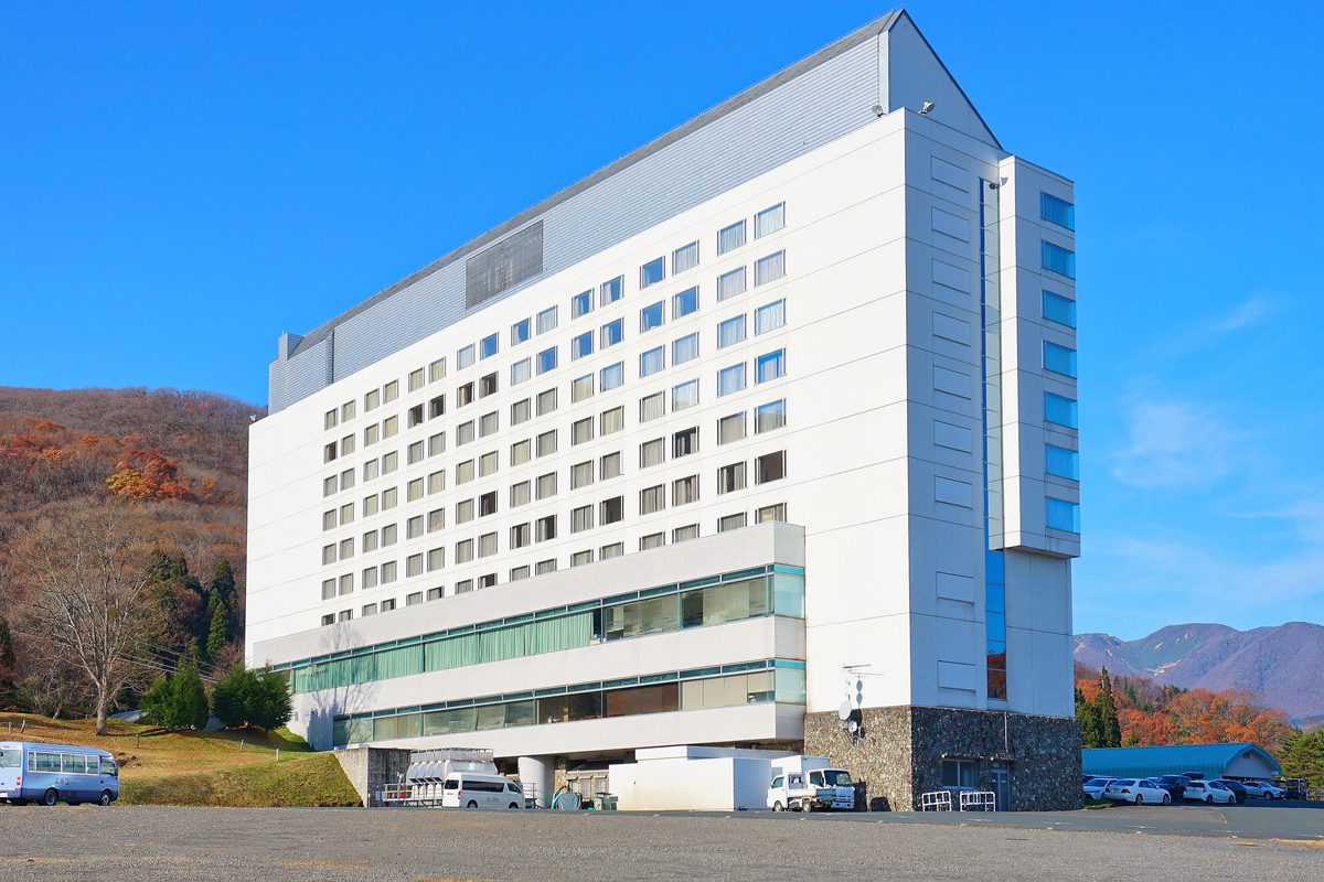 晴天下的雫石王子飯店:白色的十層樓長方形建築,屋頂一側斜面,底部是石牆基座與玻璃帷幕的低樓層;左後方是紅褐色的秋季山坡,右遠方有山脊,前面是碎石停車場,停著一台淡藍色中型巴士與一台白色廂型車