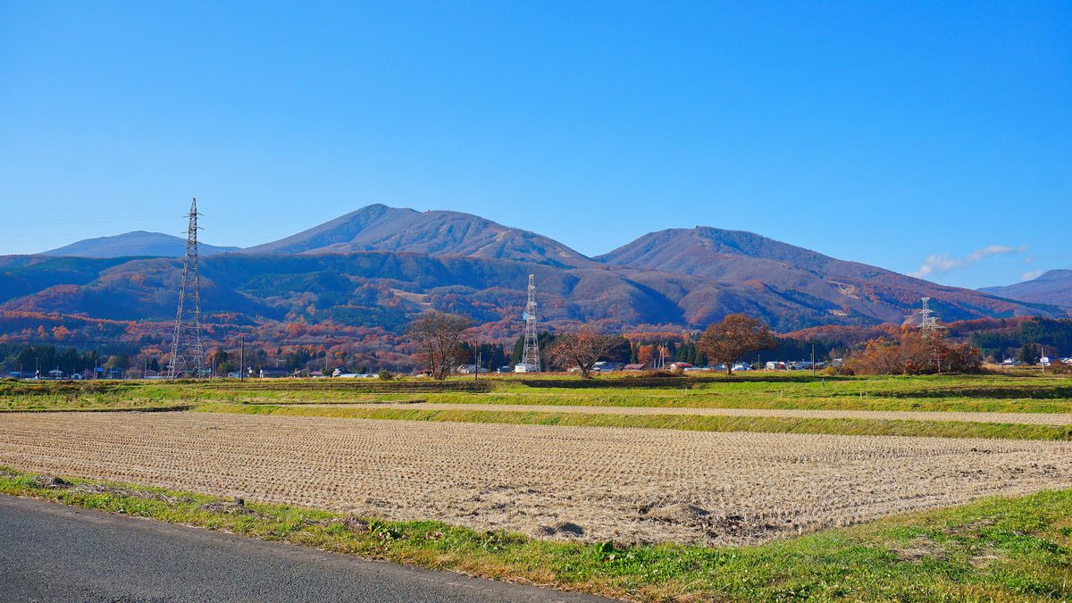從雫石町的田野望向高倉山與小高倉山:兩座紅褐色的秋季山頭並排,山腰有幾道淺色的雪道切線,前景是收割後的稻田與輸電鐵塔