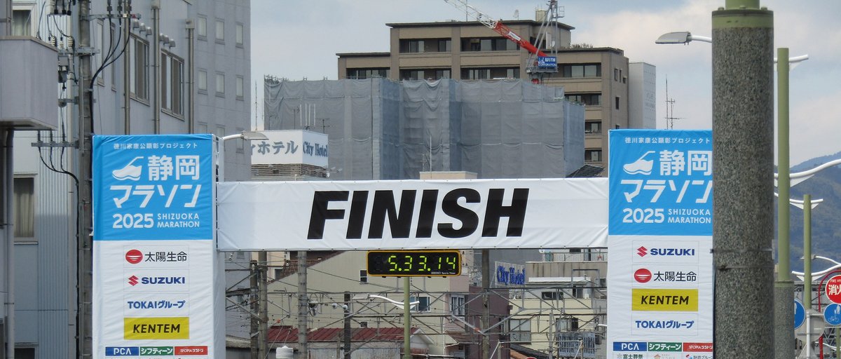 2025 年靜岡馬拉松終點:白底黑字 FINISH 的拱門橫跨道路,兩側是藍白色的「静岡マラソン 2025」贊助直幅,拱門下的計時鐘顯示 5:33:14,背後有電車架線和寫著 City Hotel 的飯店招牌
