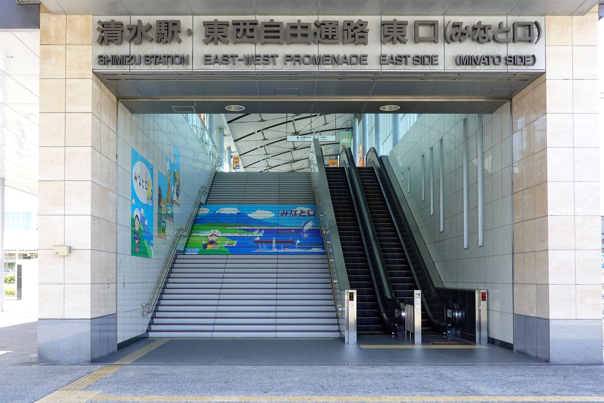 JR 清水站東西自由通路東口的入口:上方石材招牌寫著「清水駅・東西自由通路 東口(みなと口)」與英文 Shimizu Station East-West Promenade East Side,入口內是一道寬樓梯和兩座手扶梯,樓梯中段貼著港口風景的彩色壁畫