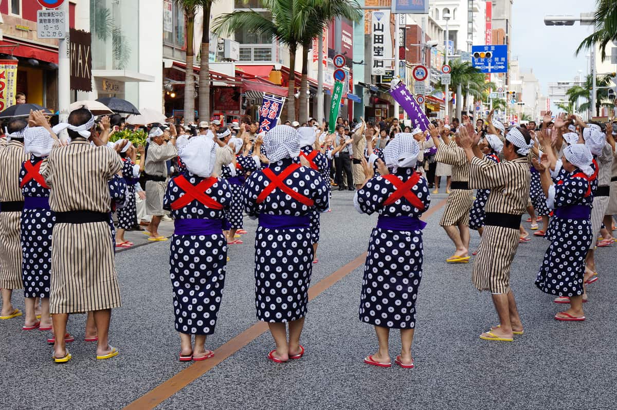 Kokusai-dori closed to traffic during the Shuri Castle Festival: a front row of dancers in navy yukata patterned with white rings, purple sashes, red cross-tied cords across their backs and white cloths bound round their heads, behind them dancers in beige-and-brown striped kimono, all with hands raised clapping, and spectators with parasols packed under the shopfront arcades, palm trees and shop signs including 山田宝石 in the background