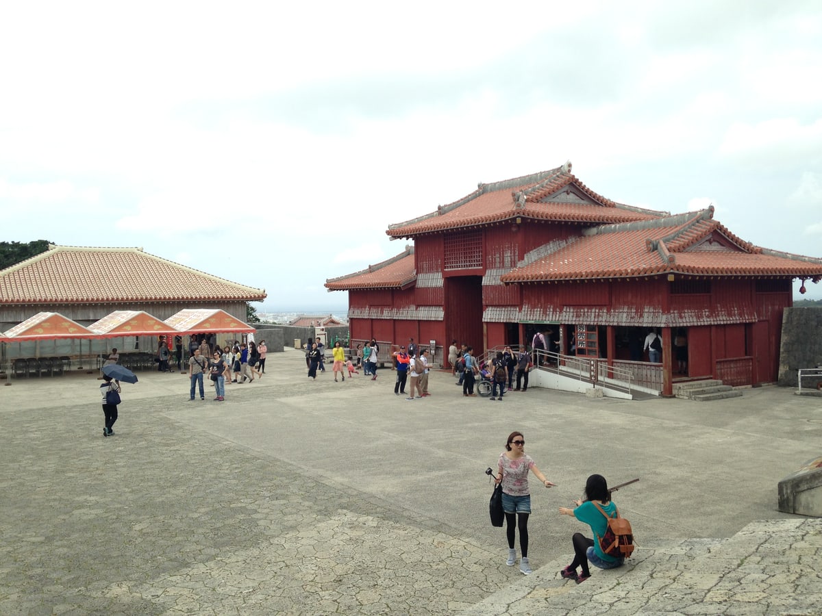 Looking back at the Kofukumon from the Hoshinmon: the broad paved Shicha-nu-Una courtyard with several dozen visitors spread across it, the vermilion Kofukumon building on the right with an open counter frontage where people are queueing and a wheelchair ramp alongside, red-and-gold parasols over rows of chairs at a low building on the left, and the Naha cityscape and sea beyond the wall in the distance