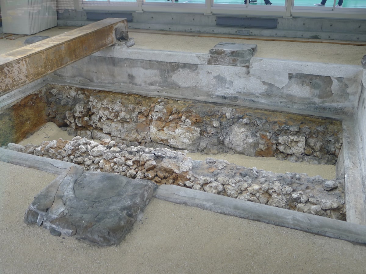 The platform remains beneath the Seiden of Shuri Castle: an excavated rectangular pit holding two parallel courses of rough coral-limestone blocks with fine sand between them, framed by grey concrete edging and a rusted steel beam, and at the very top of the frame, through glazing, the feet of visitors walking on a green floor above