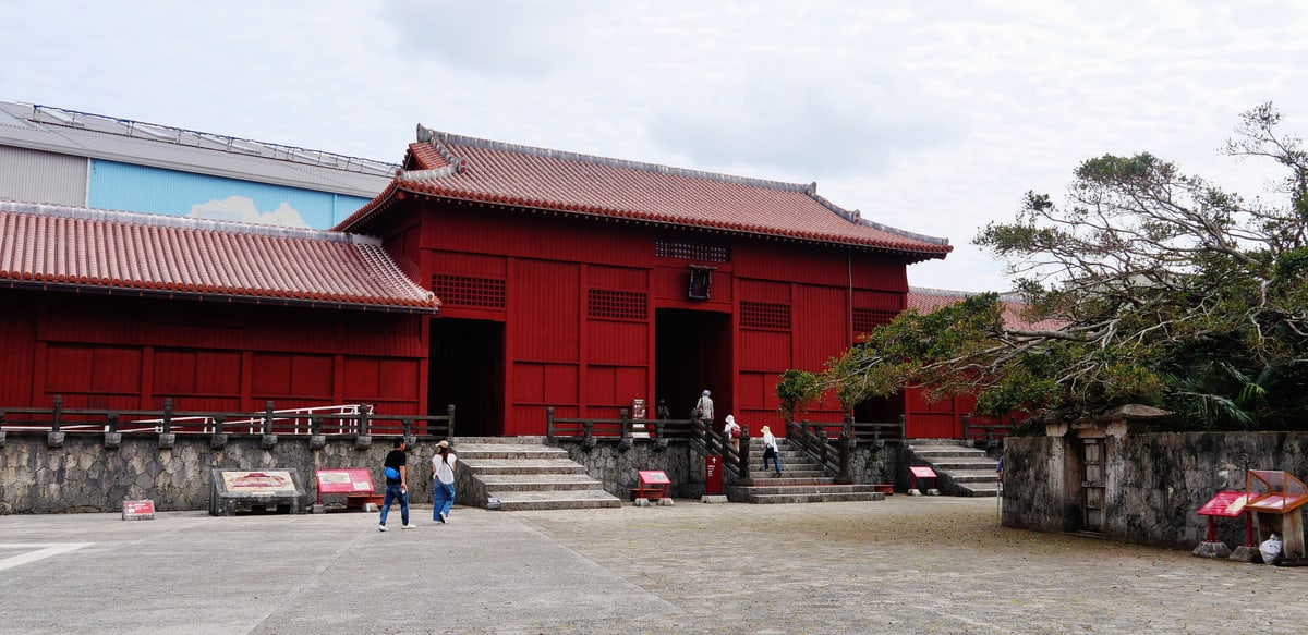 The Shicha-nu-Una at Shuri Castle: a wide grey paved courtyard with a few interpretive panels and benches set on it, facing the vermilion timber Hoshinmon gate and its low red flanking ranges, a black plaque hung above the gateway, a large tree to the right, and a big white-and-blue construction structure visible behind the roofline on the far left