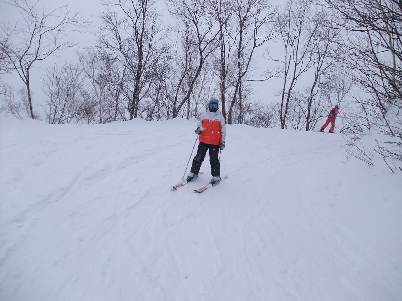 Skier coming down a piste towards the camera in a pale grey helmet and blue-lensed goggles, neck gaiter covering the lower face, white jacket with an orange chest panel, black trousers, light gloves, poles in both hands and red-and-white skis
