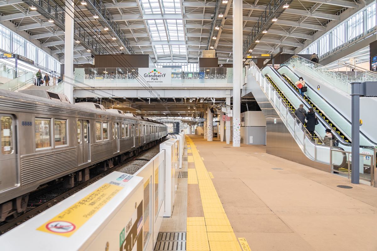 南町田グランベリーパーク站月台:左邊停著銀色的田園都市線電車,月台邊有白色月台門,中間掛著「Grandberry Park Minami-machida」的招牌,右邊是往上的電扶梯,挑高的鋼構屋頂有天窗