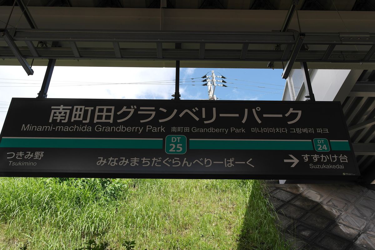 南町田グランベリーパーク站的月台站名牌:深色底白字,大字寫「南町田グランベリーパーク」,下面有英文 Minami-machida Grandberry Park 與中韓文,中間綠色車站編號 DT25,左邊前一站つきみ野、右邊下一站すずかけ台,背景是藍天與月台下方的草坡