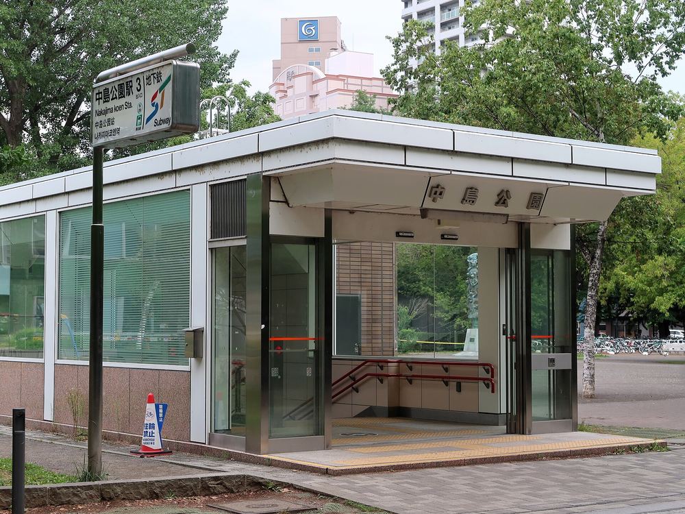 地下鐵南北線中島公園站 3 號出入口:平頂的玻璃與不鏽鋼小站房,屋簷上寫著「中島公園」,左側站牌寫「中島公園駅 3 Nakajima koen Sta.」,周圍是綠樹,夏天