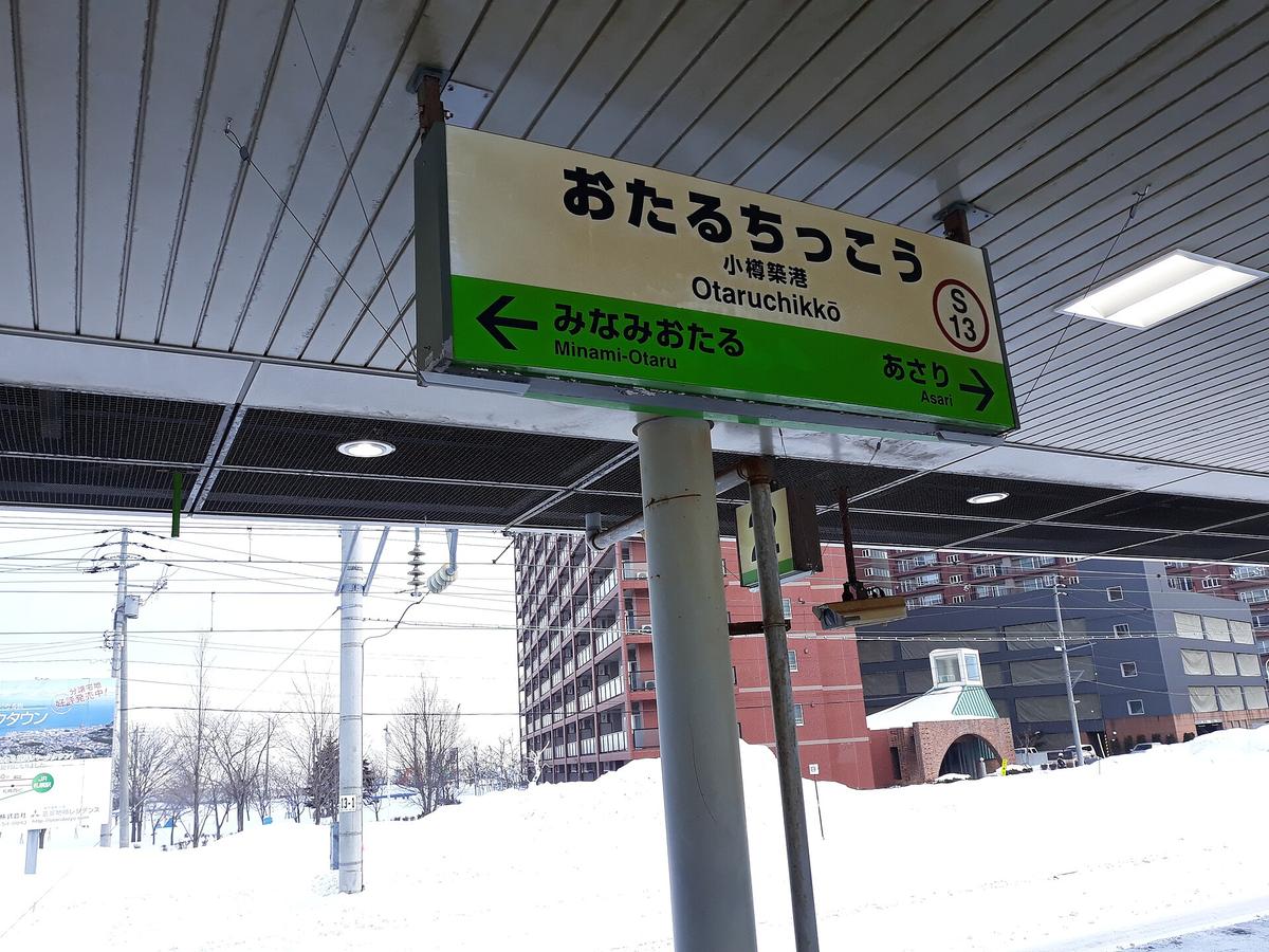 冬天的小樽築港站月台:屋簷下掛著站名牌「おたるちっこう 小樽築港 Otaruchikkō S13」,左箭頭往みなみおたる、右箭頭往あさり,月台外是厚厚的積雪與一棟紅磚色大樓