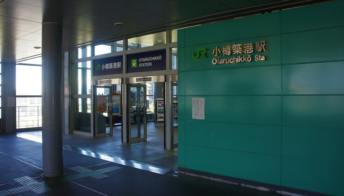 JR 小樽築港站的入口:有屋頂的通道盡頭是一排玻璃門,門上方掛著「小樽築港駅」「OTARUCHIKKŌ STATION」兩塊站名牌,右側青綠色牆面寫著「JR 小樽築港駅 Otaruchikkō Sta」