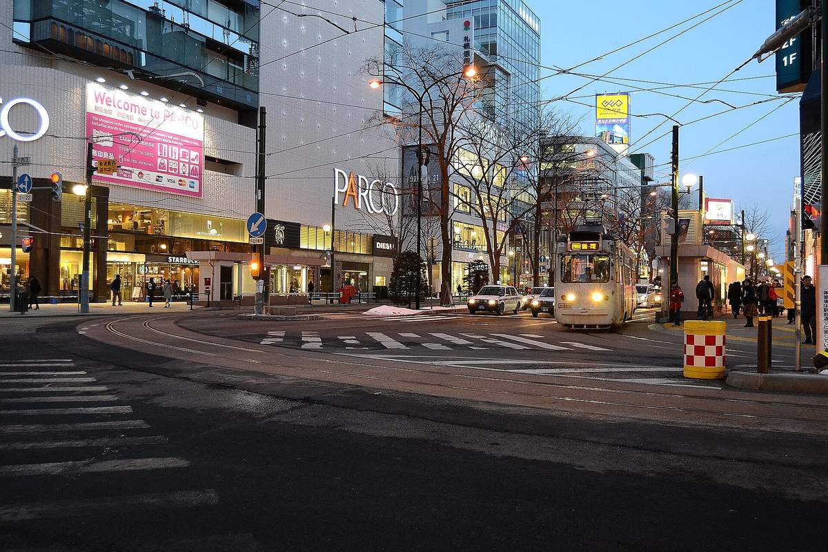 傍晚的札幌站前通:一台白色車身的一般市電亮著車頭燈沿彎道開過來,左側是 PARCO 百貨與 Starbucks,路邊殘雪,行道樹沒有葉子