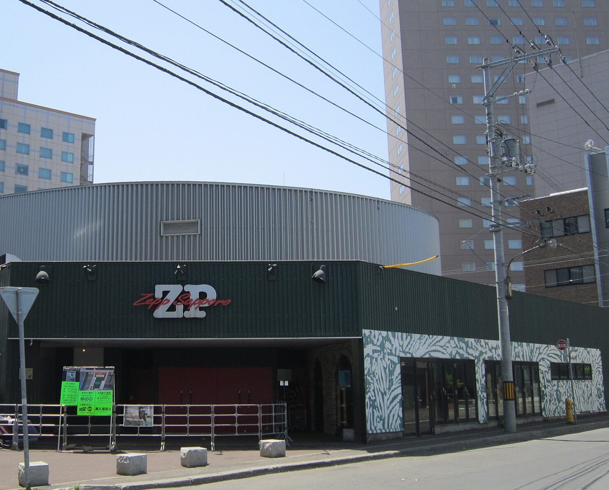 演唱會場地 Zepp Sapporo 外觀:深綠色鐵皮外牆上紅白色「ZP Zepp Sapporo」標誌,後方是銀色圓筒形的場館本體,入口前一排鐵柵欄,背後一棟高樓飯店,晴天