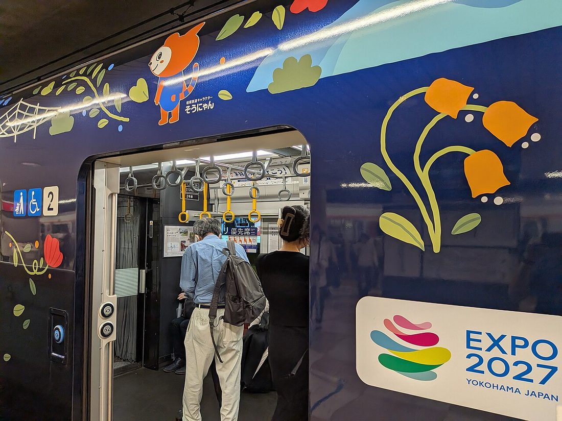 A Sotetsu train in navy livery wrapped with orange bell-shaped flowers, green leaves, a red bloom and a spiderweb motif; an orange cat character labelled Sounyan sits above the doorway and a white rounded plaque carries the colourful EXPO 2027 YOKOHAMA JAPAN logo, with two passengers boarding through the open door
