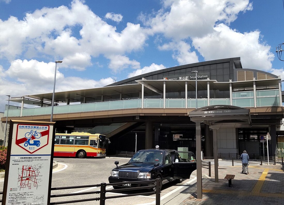 The elevated station building of Seya Station on the Sotetsu Main Line, with a sign reading SOTETSU 瀬谷駅 Seya Station, a taxi rank shelter with a black taxi below it, a yellow-and-red route bus, and a Yokohama City no-bicycle-parking noticeboard in the foreground
