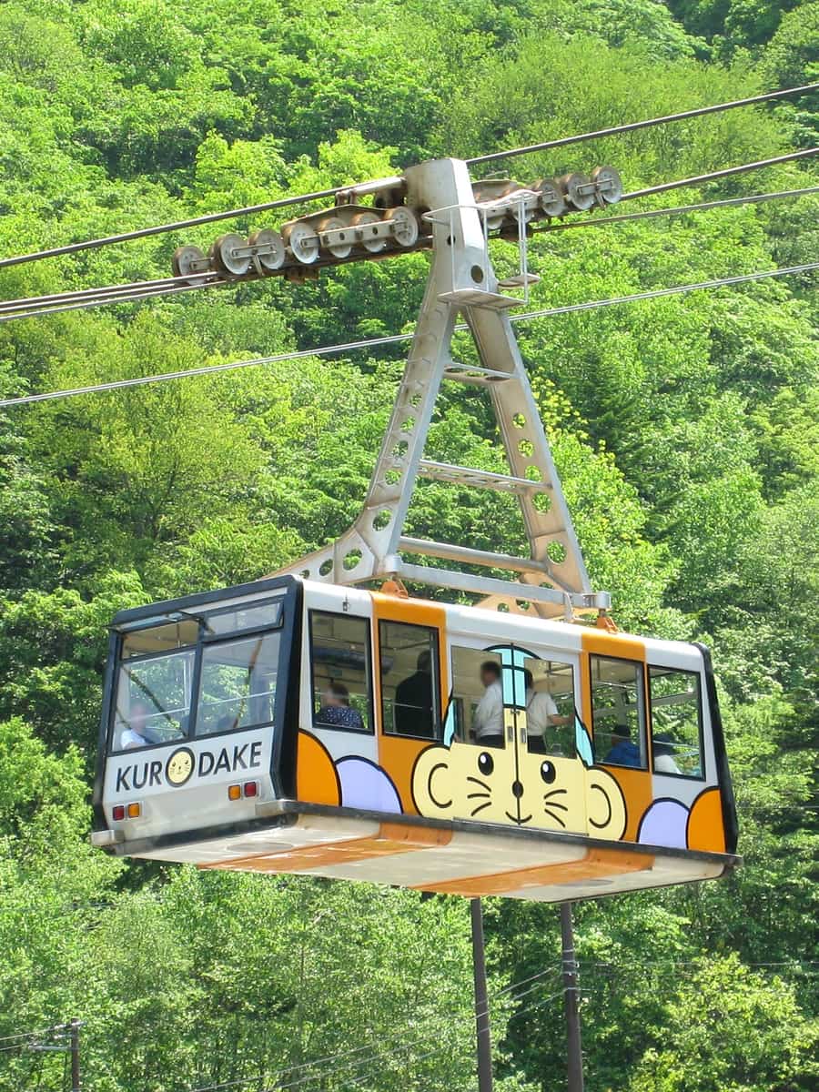 Kurodake Ropeway gondola with KURODAKE lettering and a mascot design, travelling above green forest
