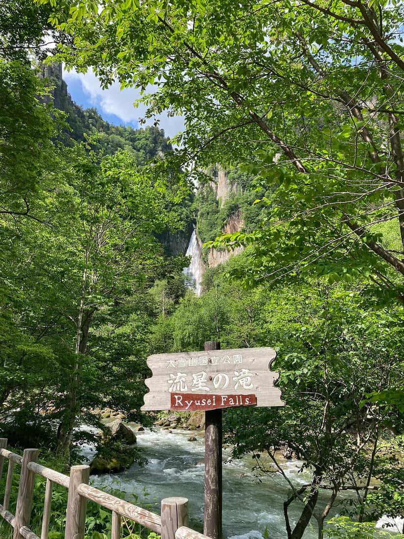 Wooden Ryusei Falls signpost with the waterfall dropping from the clifftop behind and the Ishikari River in front