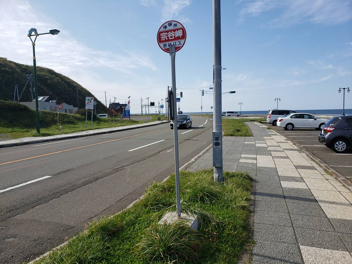 宗谷岬路邊的圓形紅白站牌,寫著「停留所 宗谷岬 宗谷バス」,立在人行道旁的草地上,左邊是國道,右邊是停車場和遠處的海,晴天