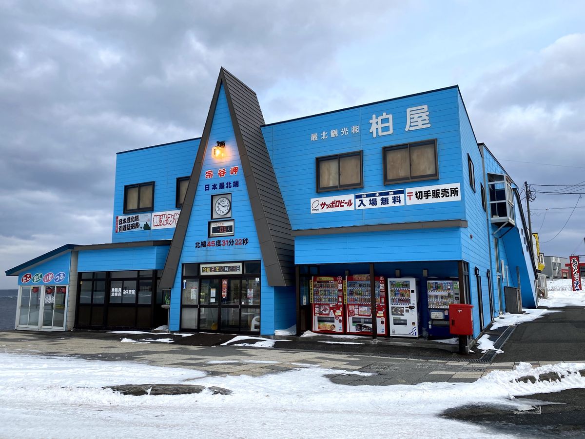 12 月積雪的宗谷岬,藍色兩層樓的柏屋土產店,三角形屋頂下寫著「宗谷岬 日本最北端」,右側招牌「最北観光株 柏屋」「流氷館 入場無料」「切手販売所」,左翼二樓牆上掛著「日本最北端到着証明書」的看板,門前一排自動販賣機
