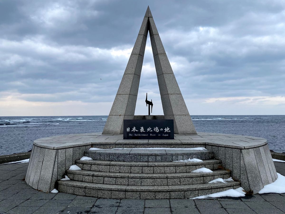 近看「日本最北端の地」碑:兩片花崗岩斜板合成三角形,中間立著一座金屬雕塑,黑色碑板寫著「日本最北端の地 The Northernmost Point in Japan」,台階邊緣有殘雪,背後是起浪的海和厚雲
