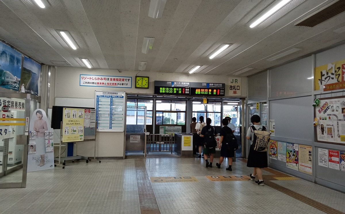 The ticket gate at JR Goshogawara Station, where an attendant stands at the barrier checking tickets by hand as several passengers, seen from behind, file through beneath a two-line departure board