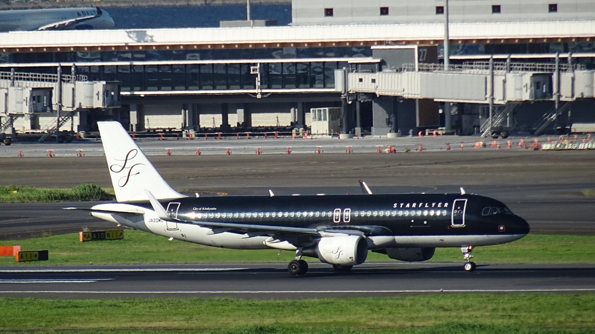 An all-black StarFlyer Airbus A320 taxiing at Haneda, STARFLYER titles in white on the forward fuselage, a white tail carrying a black SF monogram, registration JA20MC near the rear, terminal jet bridges behind