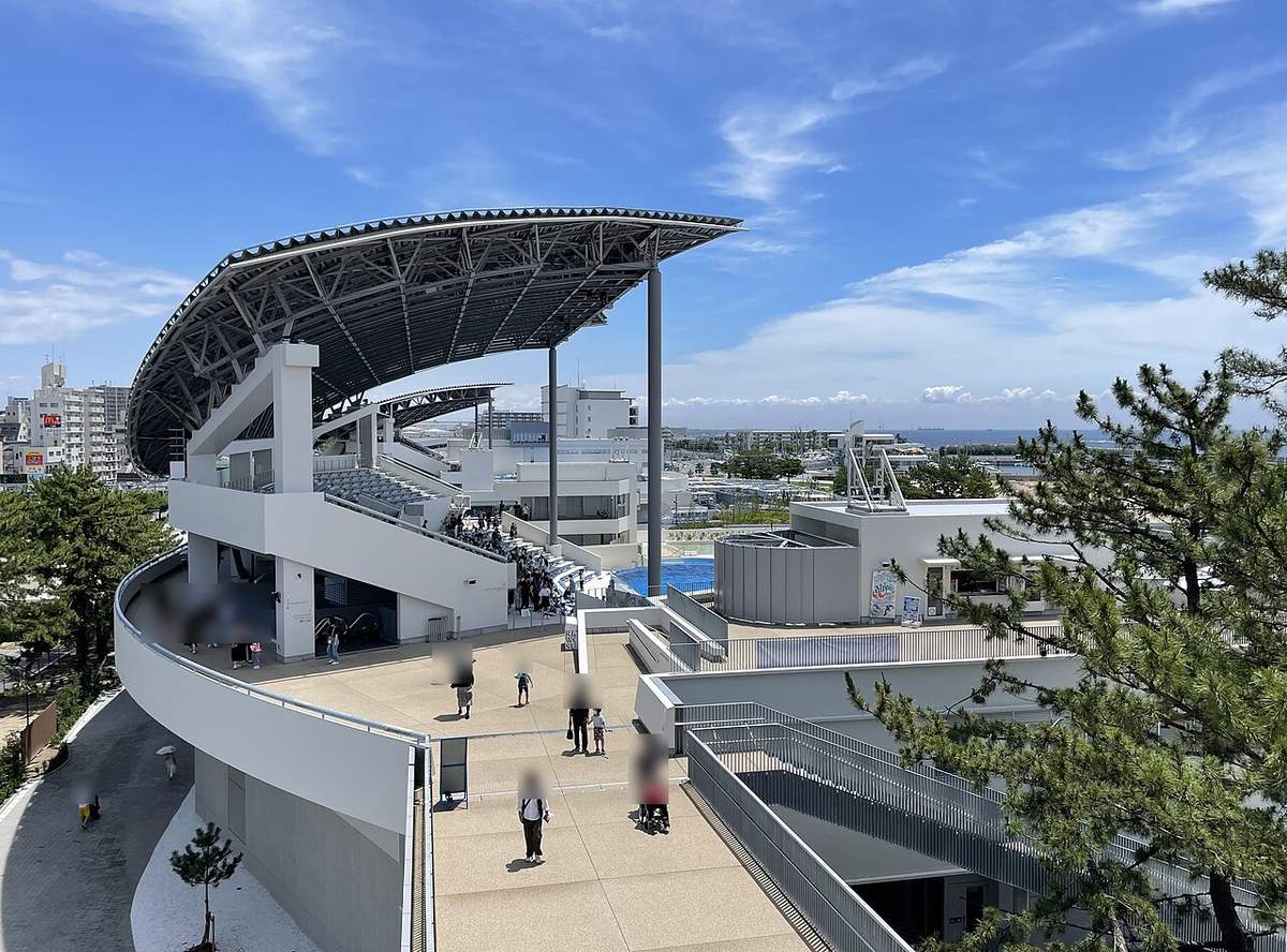 從高處步道看海豚表演場ドルフィンスタディアム,黑色鋼構弧形大屋頂下是白色階梯看台,看台前方露出一角藍色表演池,遠處是住宅大樓與海