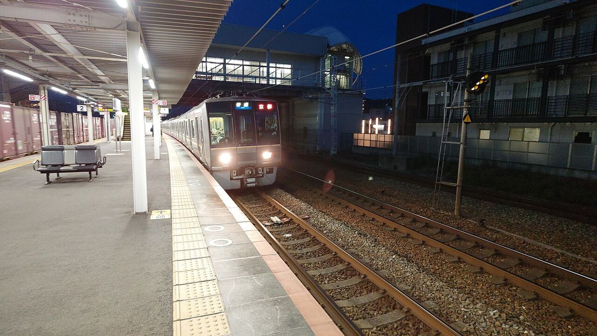 傍晚的 JR 須磨海浜公園站月台,一列銀色車身、亮著頭燈的 JR 西日本 207 系電車正駛進月台,左側月台有長椅與 2 號乘車位置標示,後方是車站跨線通道